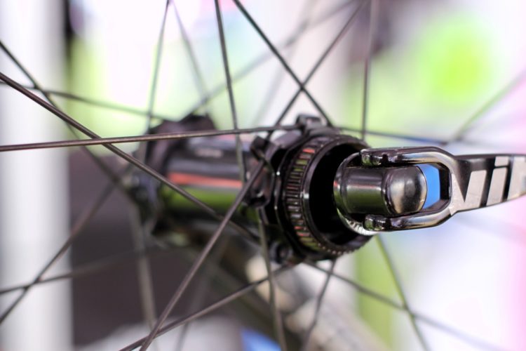 Close-up view of a bicycle wheel hub and spokes, showcasing the intricate design and metallic elements of the hub and the tensioned spokes radiating outward. The background features blurred colors, indicating an outdoor or cycling event setting.