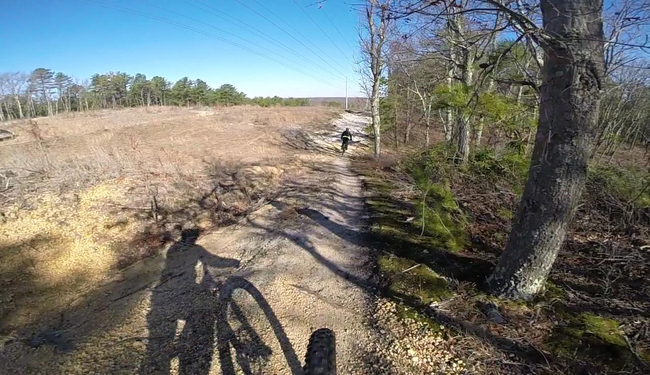 A mountain biker riding along a gravel path surrounded by trees and open fields under a clear blue sky. The foreground features the shadow of the bike, and in the background, another cyclist is visible on the trail. Allaire State Park mountain bike trail.