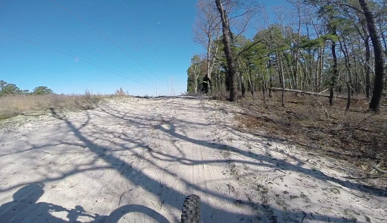 Mountain biking trail through a sandy path surrounded by trees, with sunlight filtering through branches and power lines visible in the background. The perspective shows the cyclist's shadow and bike tire in the foreground, creating a sense of motion. Allaire State Park mountain bike trail.