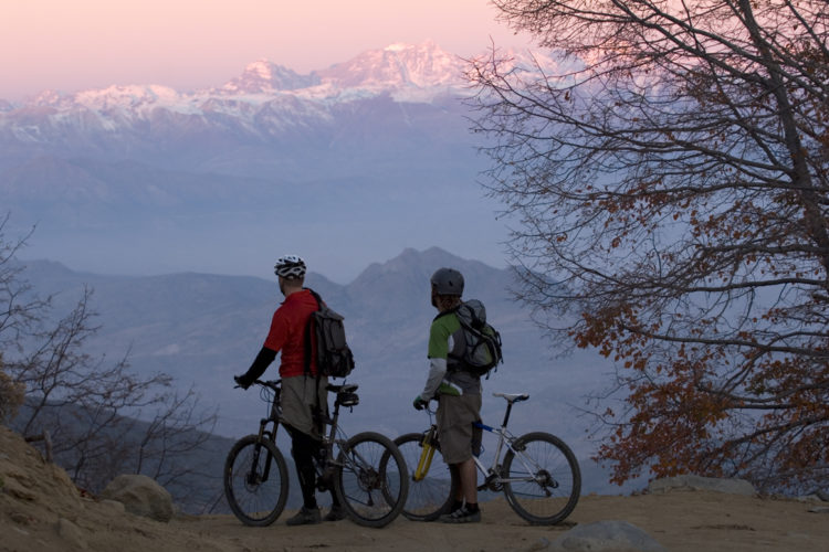 Two mountain bikers stand on a hillside, looking out over a scenic landscape of distant snow-capped mountains at dawn. One biker wears a red shirt and a helmet, while the other is dressed in a green shirt with a gray jacket. Both have backpacks and their bikes beside them. The sky is tinged with soft pink hues, and trees with sparse leaves frame the scene.