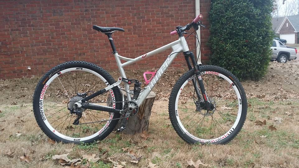 Niner Air 9: Alt text: A mountain bike with a silver and black frame is resting against a tree stump in a grassy area, with a brick wall in the background. The bike features white wheels and pink accents. Fallen leaves are scattered on the ground, and a parked vehicle is visible in the distance.