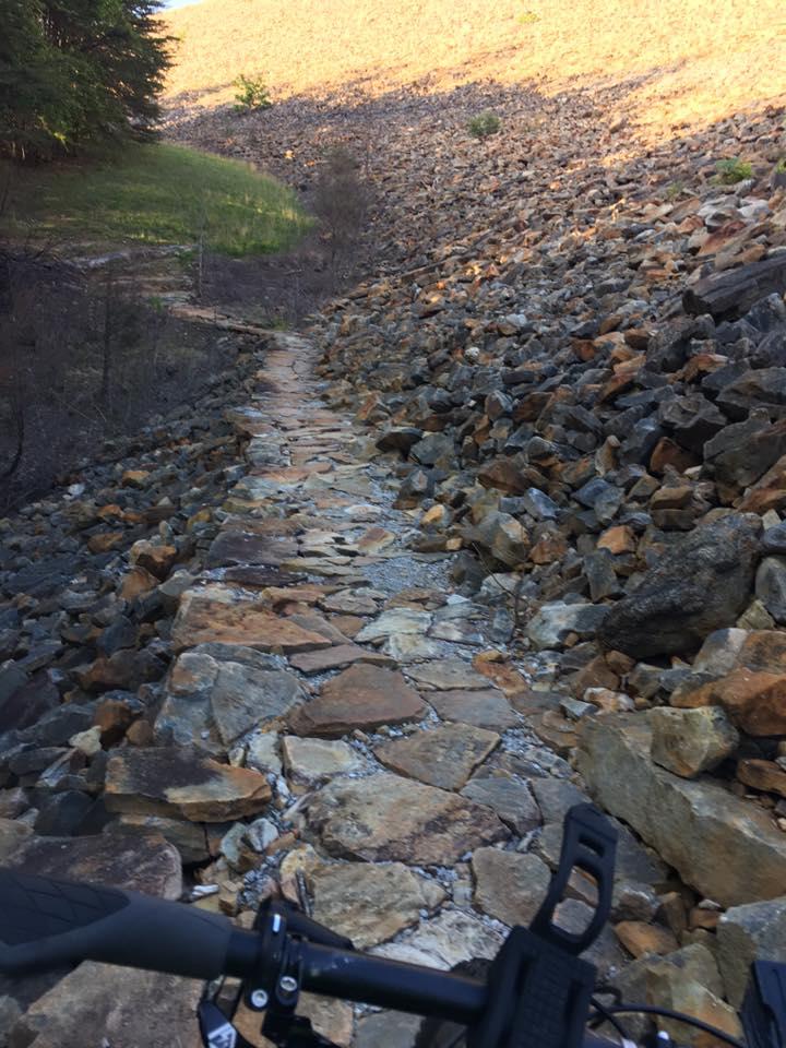 A rocky pathway winding through a hillside, with large stone slabs forming a narrow trail. The path is flanked by loose rocks and sparse vegetation on either side, leading up to a grassy area in the distance. The foreground shows the handlebars of a bicycle, suggesting that the image was taken while riding. Raccoon Mountain Trail Network mountain bike trail.