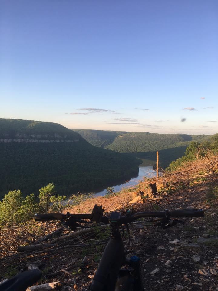 A scenic view of a river winding through lush green hills, captured from a high vantage point. A mountain bike is positioned in the foreground, resting on rocky terrain. The sky is clear with soft clouds, indicating a tranquil evening. Raccoon Mountain Trail Network mountain bike trail.