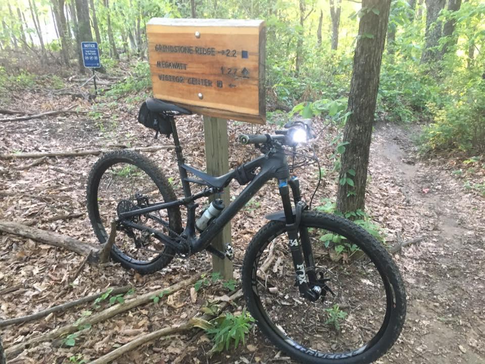 Specialized Camber Expert Carbon 29: A mountain bike parked near a trail sign in a wooded area. The sign indicates directions to various trails, including "Grindstone Ridge" and "Megawatt," along with distances to the "Visitor Center." Surrounding the bike and sign are lush greenery and leaves on the ground, suggesting a natural outdoor setting.