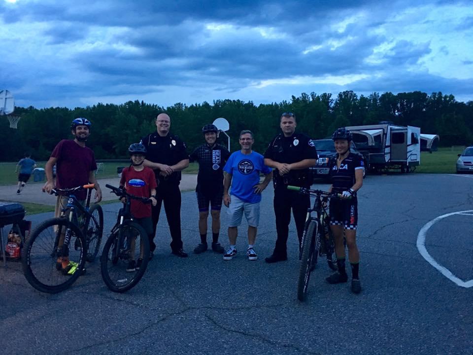 A group of six individuals stands together in a park setting, with bicycles beside them. The group includes four adults and two children, one of whom is wearing a red shirt and another a blue shirt. Two of the adults are police officers in uniforms. The background features a basketball hoop and a recreational vehicle, under a cloudy sky. Farris Park mountain bike trail.