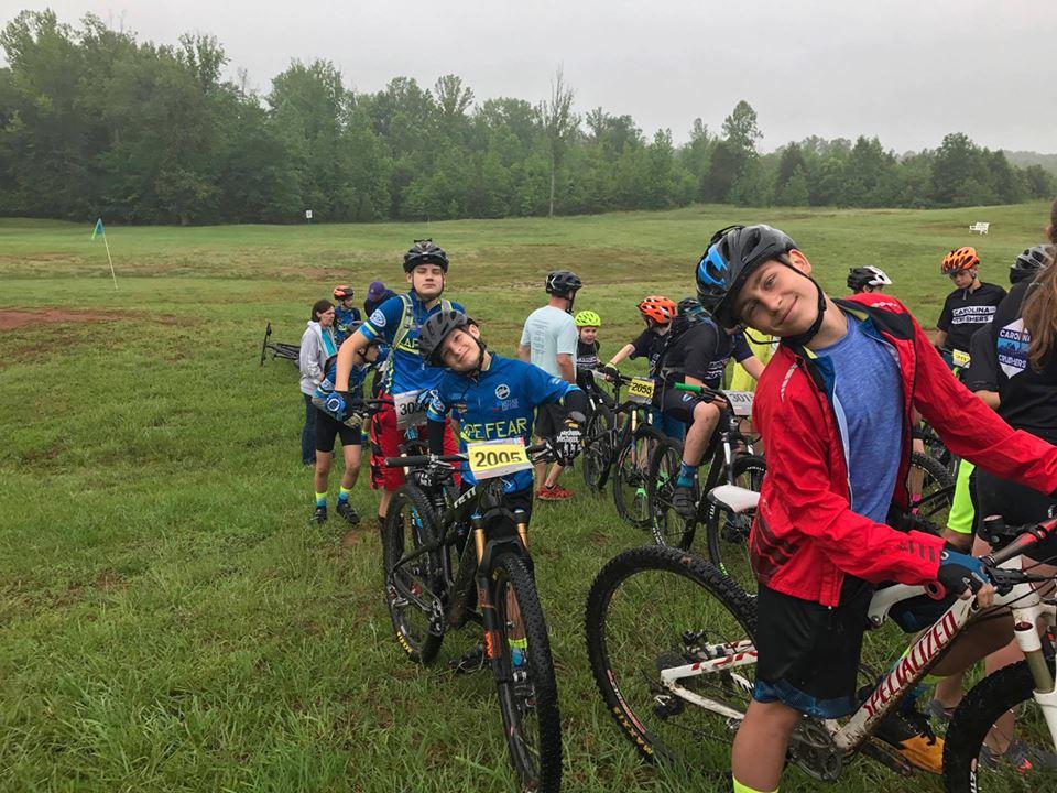 A group of kids gathered on a grassy field preparing for a mountain biking event. They are wearing colorful cycling jerseys and helmets, standing beside their bicycles. Some children are smiling and posing for the camera, with a backdrop of trees and a cloudy sky. Farris Park mountain bike trail.