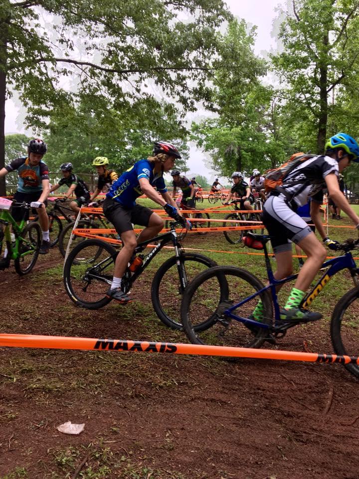 A group of cyclists navigate a wooded course during a mountain biking event, with several riders in colorful jerseys and helmets riding their bikes alongside orange safety tape marking the trail. The scene is set in a green, grassy area, indicating an outdoor competition. Farris Park mountain bike trail.