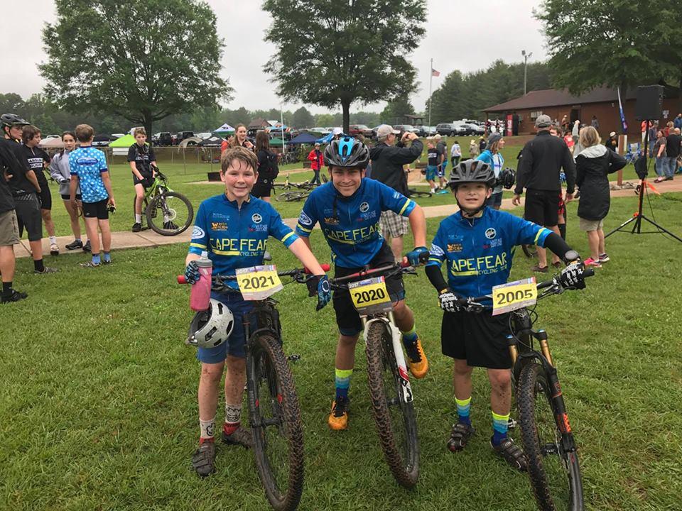Three young boys, dressed in blue and black cycling jerseys, pose with their mountain bikes at a grassy event area. Each boy has a race number attached to their bikes: 2021, 2020, and 2005. They are smiling and wearing helmets, with other participants and trees visible in the background, indicating a lively cycling event. Farris Park mountain bike trail.