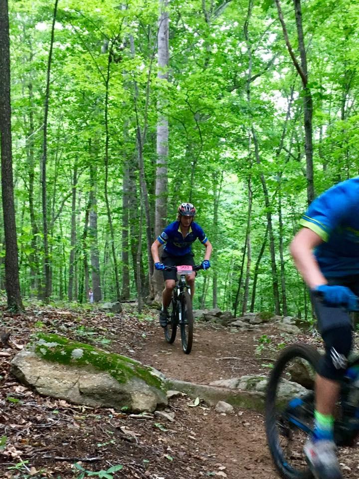 A mountain biker in a blue jersey navigates a dirt trail through a lush green forest, with rocks and foliage lining the path. Another biker can be seen partially blurred in the foreground, emphasizing the action of the ride. Farris Park mountain bike trail.