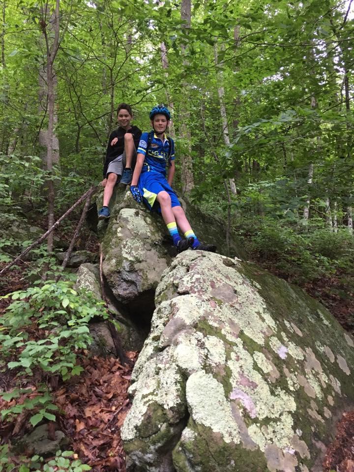 Two children sitting on a large moss-covered rock in a lush green forest. One child is wearing a bicycle helmet and colorful biking attire, while the other is dressed in a black hoodie and shorts. They are smiling and enjoying their time outdoors amidst trees and foliage. Farris Park mountain bike trail.