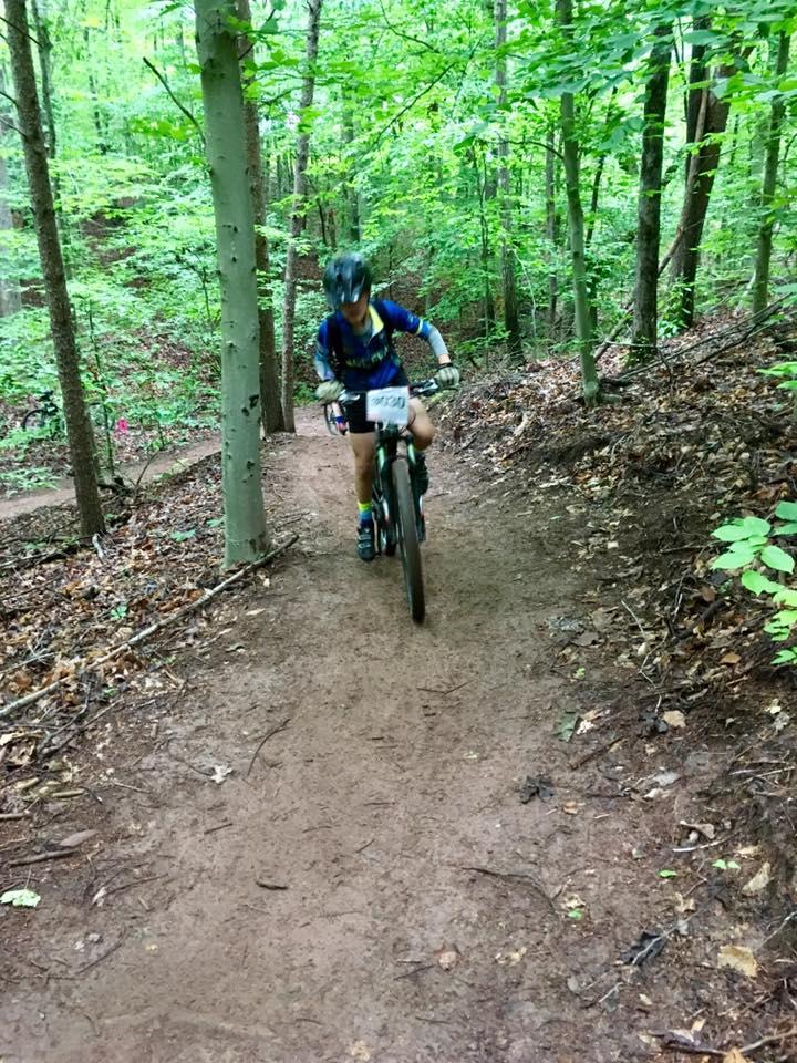 A mountain biker navigating a narrow dirt trail in a lush green forest, with trees lining the path and sunlight filtering through the foliage. The biker is wearing a helmet and sports attire, focused on the ride. Farris Park mountain bike trail.