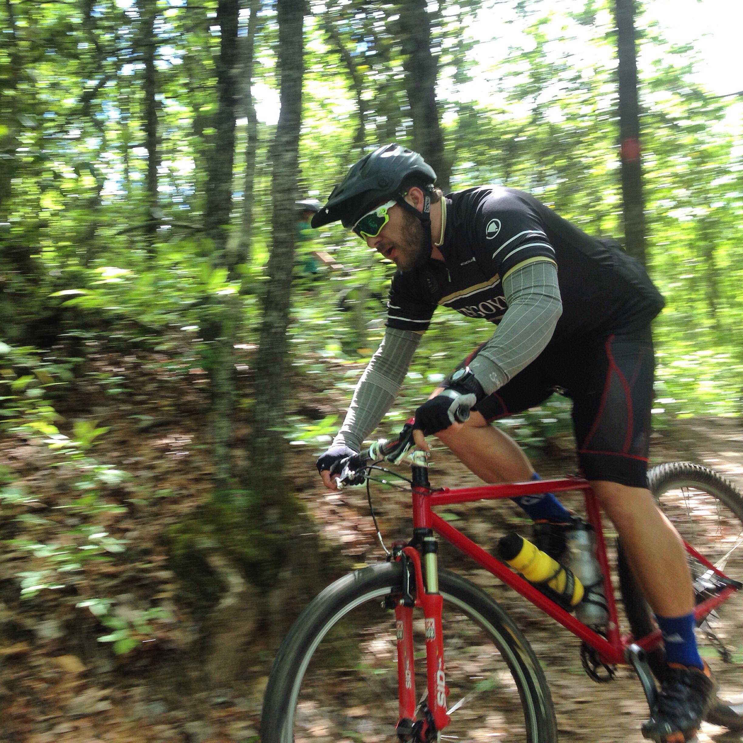 A mountain biker in motion riding along a forest trail. The cyclist is wearing a helmet and sunglasses, and is on a red bike. Surrounding greenery and sunlight filtering through the trees suggest a vibrant outdoor environment. Bracken Preserve mountain bike trail.