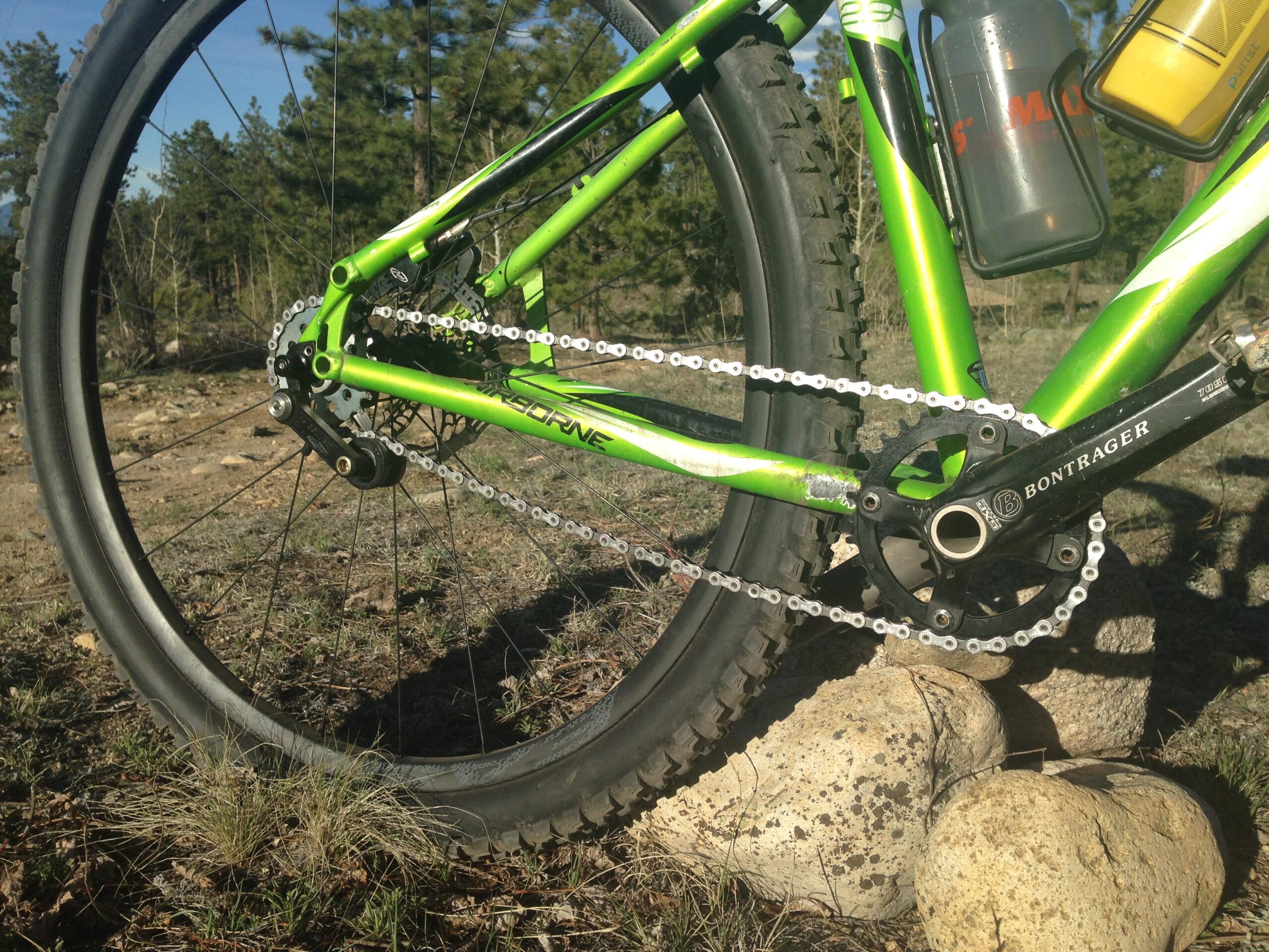 Airborne Goblin: Close-up view of a green mountain bike's drivetrain, showcasing the chain, crankset, and rear wheel on a rocky, natural terrain. The bike is equipped with a water bottle and has visible wear on the frame, indicating outdoor use in a mountainous environment.