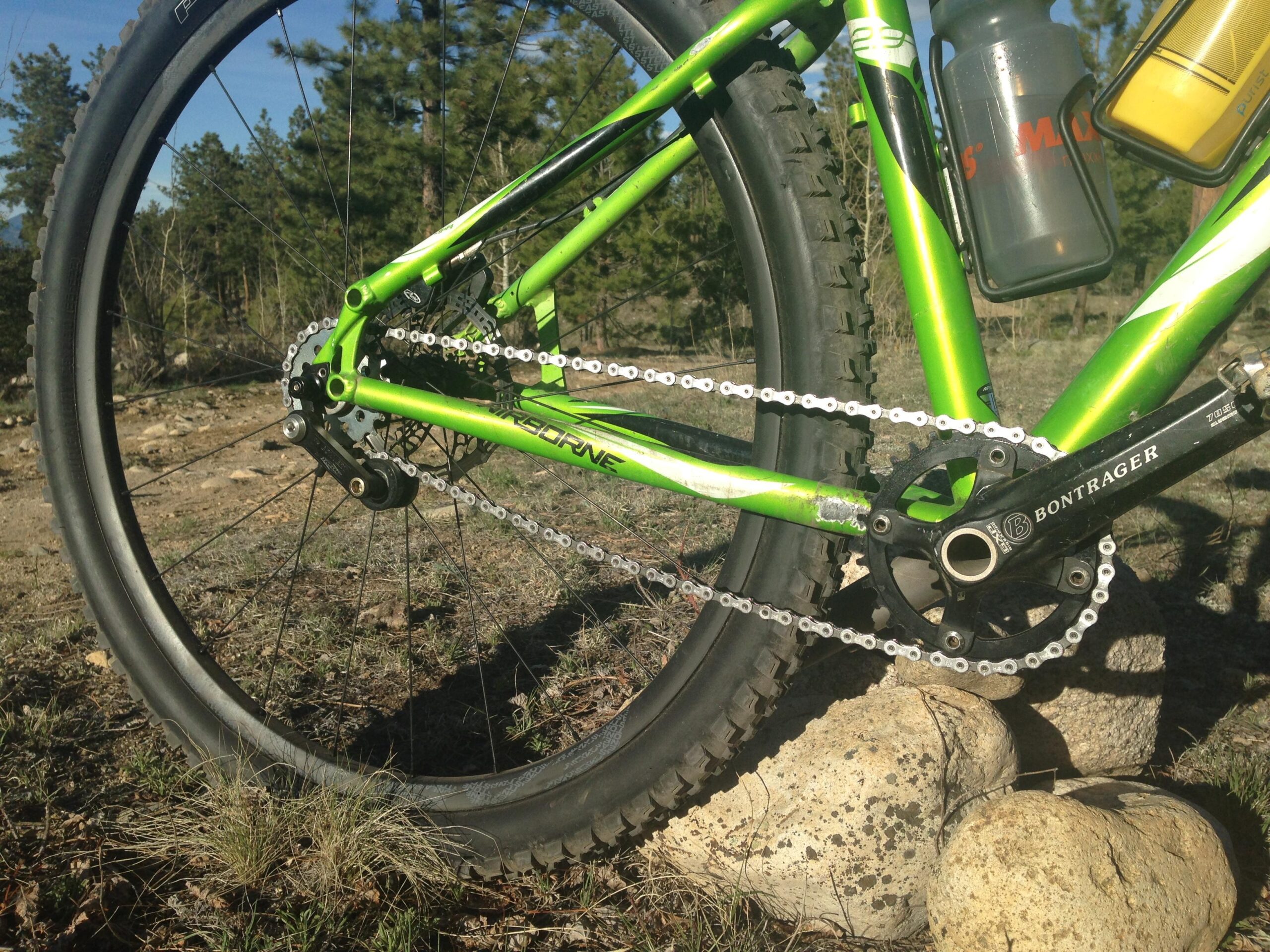 Airborne Goblin: A close-up view of a green mountain bike's drivetrain and rear wheel set against a natural landscape. The bike features a Bontrager crankset, chain, and a rear derailleur. The background includes a mix of trees and rocks, with sunlight highlighting the bike's components.