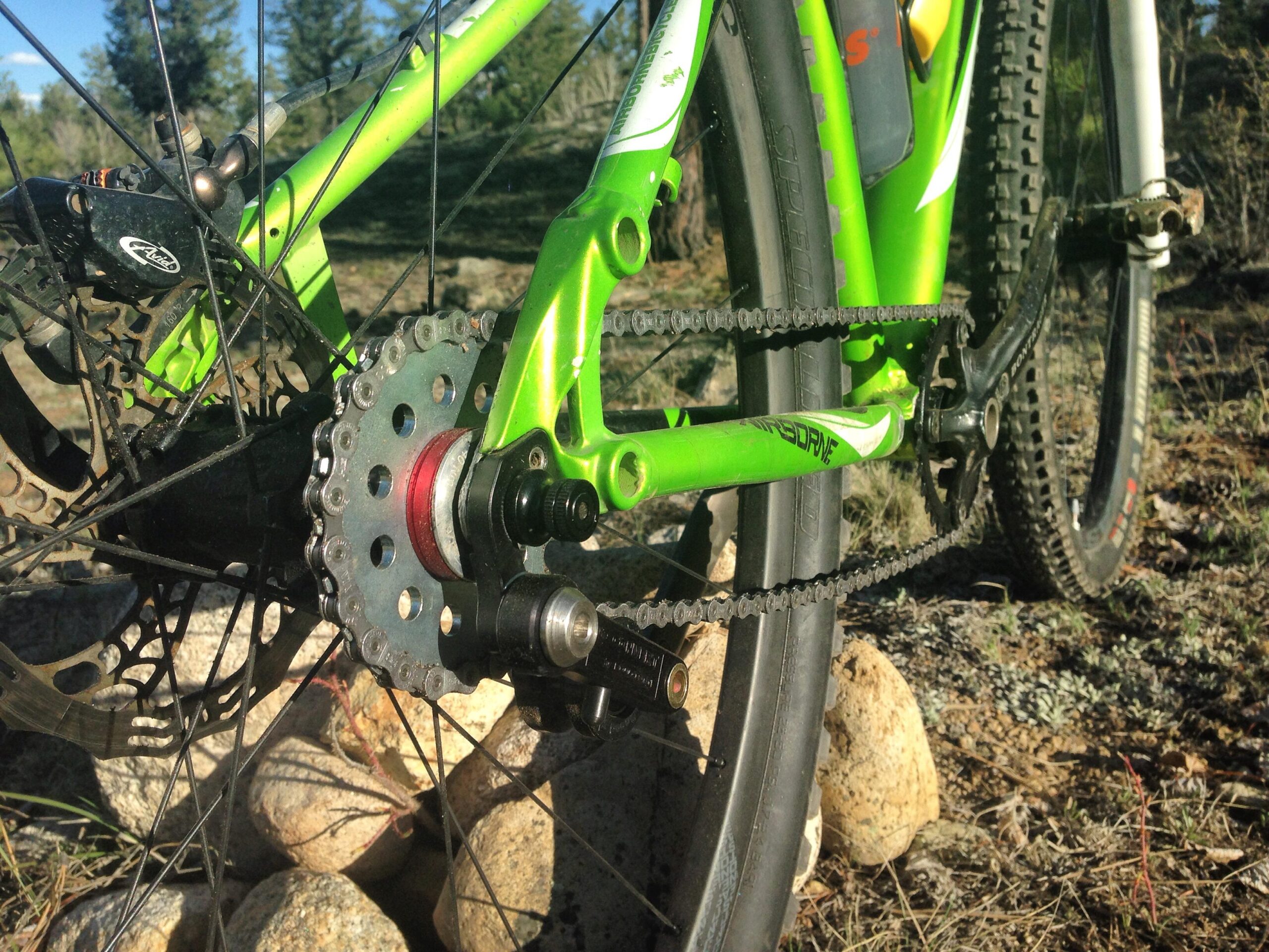 Airborne Goblin: Close-up view of the rear drivetrain of a green mountain bike, showcasing the cassette, chain, and rear derailleur against a natural background of rocks and grass.