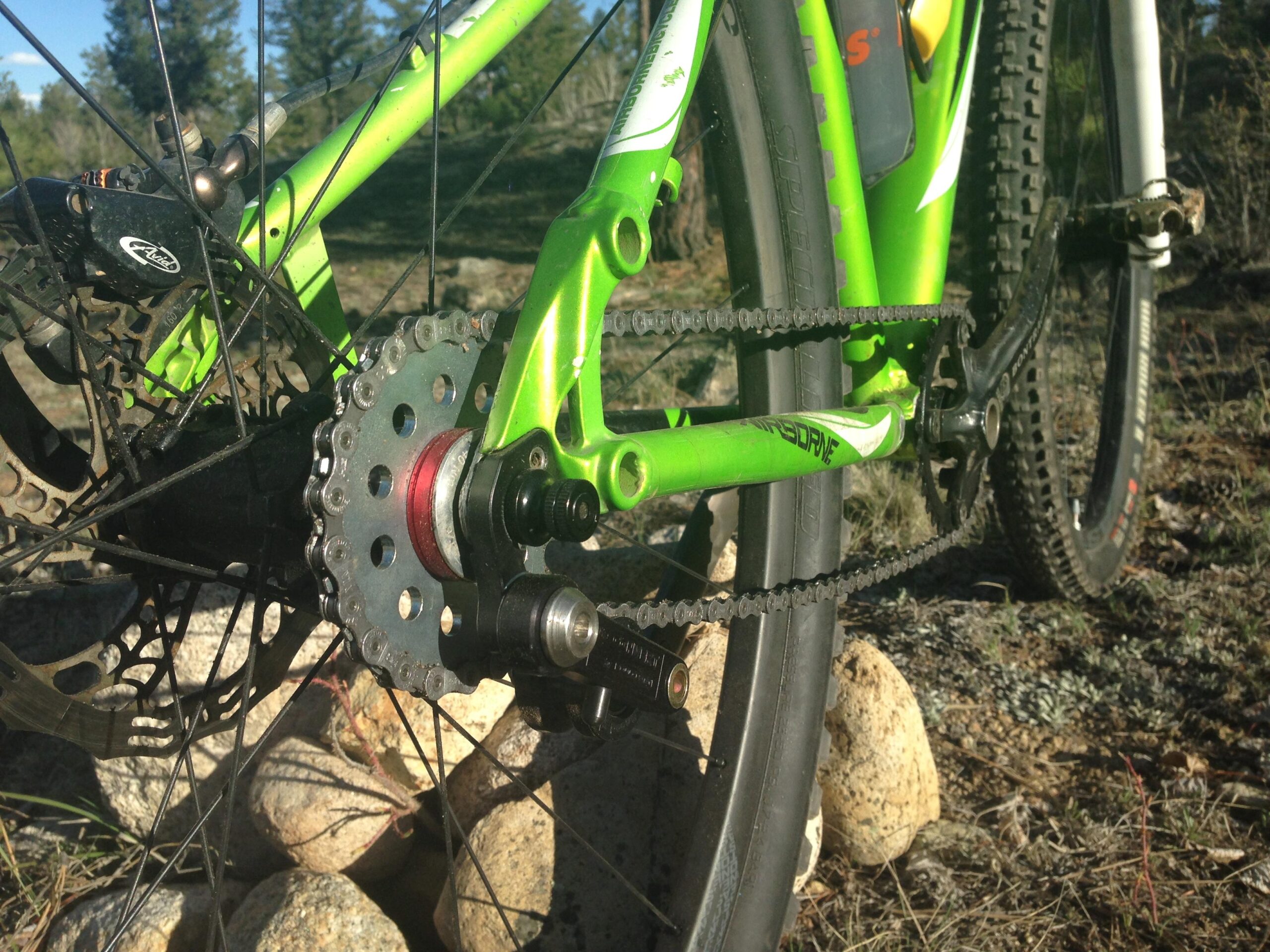 Airborne Goblin: Close-up of a green mountain bike's drivetrain, showcasing the rear derailleur, cassette, and chain. The bike is positioned on rocky terrain with trees in the background, highlighting the outdoor environment.