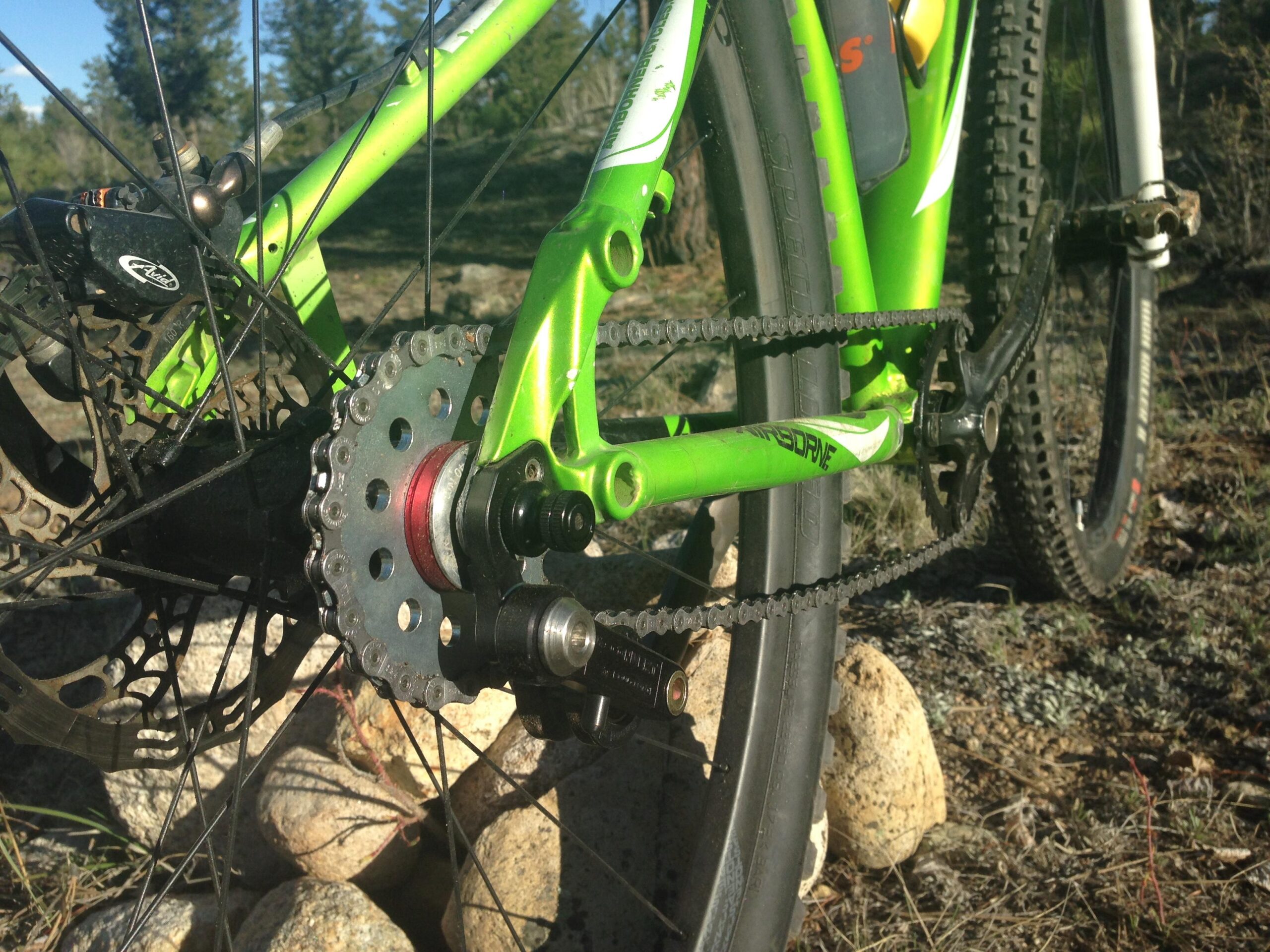 Airborne Goblin: Image of a green mountain bike, focusing on the rear drivetrain. The image shows the bike's chain, cassette, and derailleur, set against a background of natural terrain with rocks and grass.
