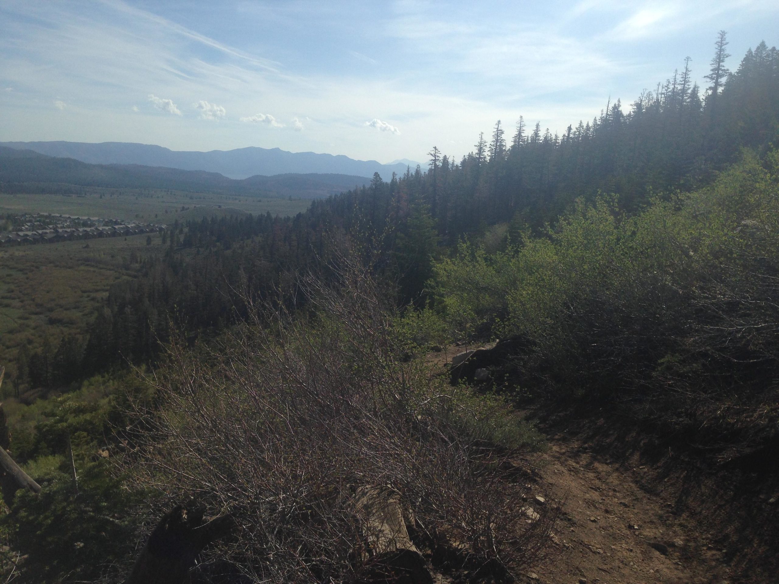 A scenic view from a hillside showing a winding dirt path leading through lush greenery, with a backdrop of mountains and a valley below. The sky is partly cloudy, and a cluster of houses is visible in the distance among the trees. Mammoth Rock Trail mountain bike trail.