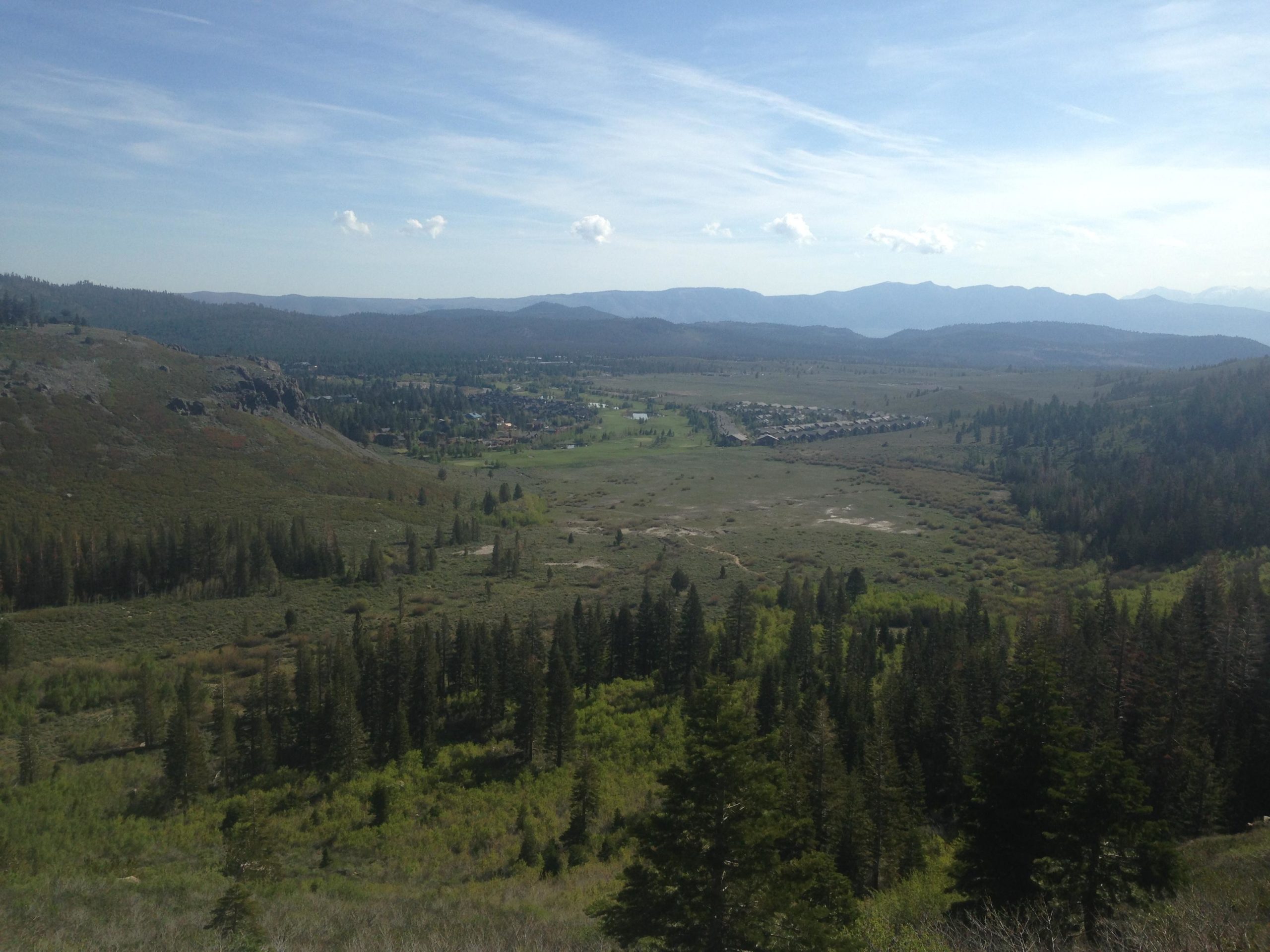 A panoramic view of a lush valley surrounded by mountains, featuring a mix of tall pine trees and open meadows. The landscape is dotted with a cluster of cabins or buildings in the distance, under a clear blue sky with a few white clouds scattered throughout. Mammoth Rock Trail mountain bike trail.