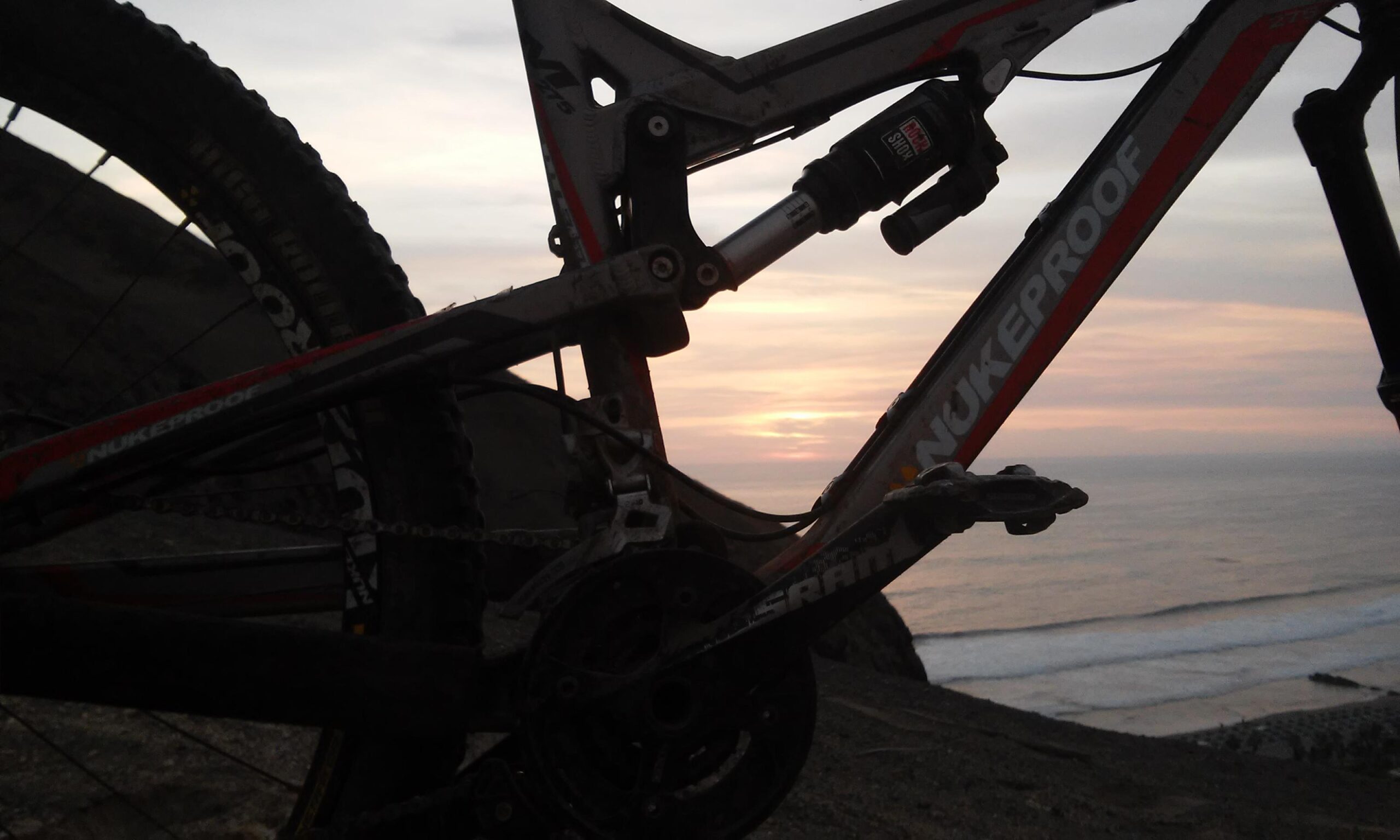 Nukeproof Mega AM 275 Comp: A mountain bike with a Nukeproof frame is silhouetted against a sunset over the ocean, with waves visible in the foreground and gentle hills in the background.