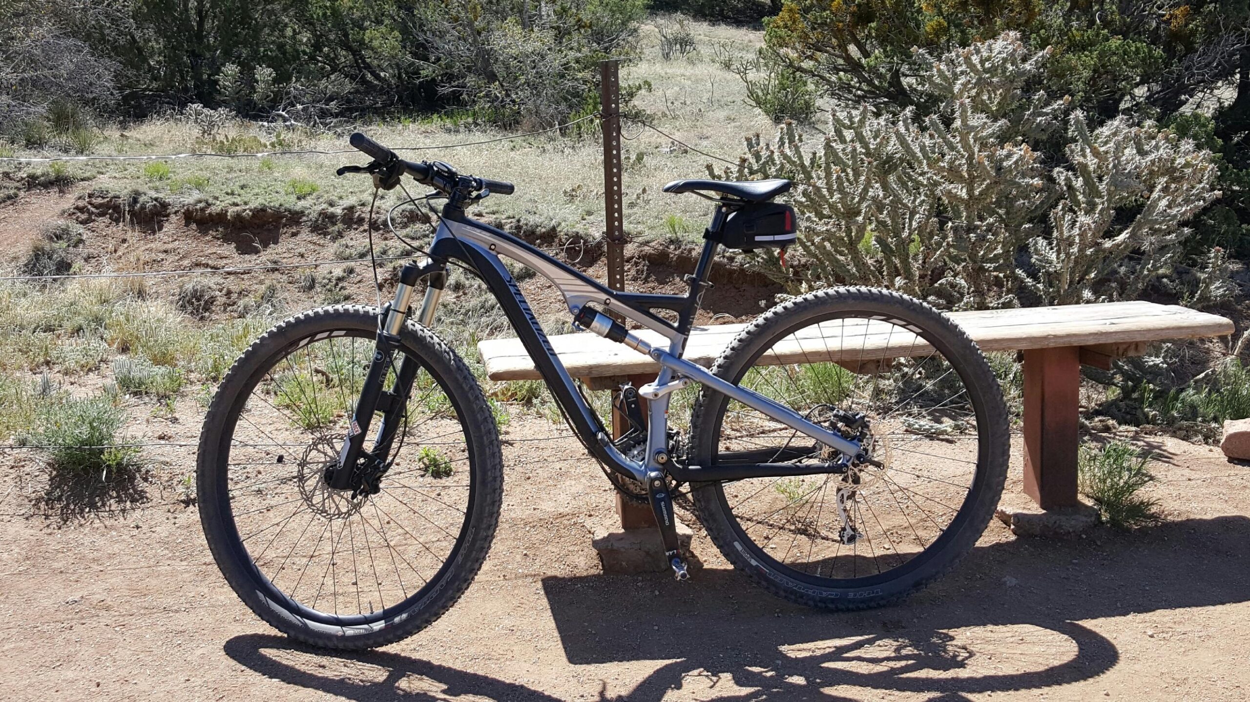 Specialized Camber Comp 29: A close-up view of a mountain bike parked next to a wooden bench on a dirt trail. The background features a mix of grass and shrubs under a clear sky. The bike has a silver and black frame with knobby tires suitable for off-road riding.