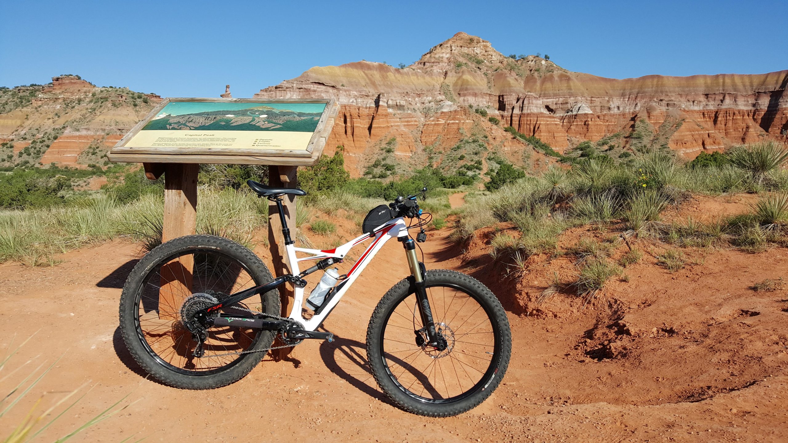 A mountain bike resting on a dirt path, next to an informational sign about Capitol Peak. The background features layered red rock formations and green vegetation under a clear blue sky. Palo Duro Canyon mountain bike trail.
