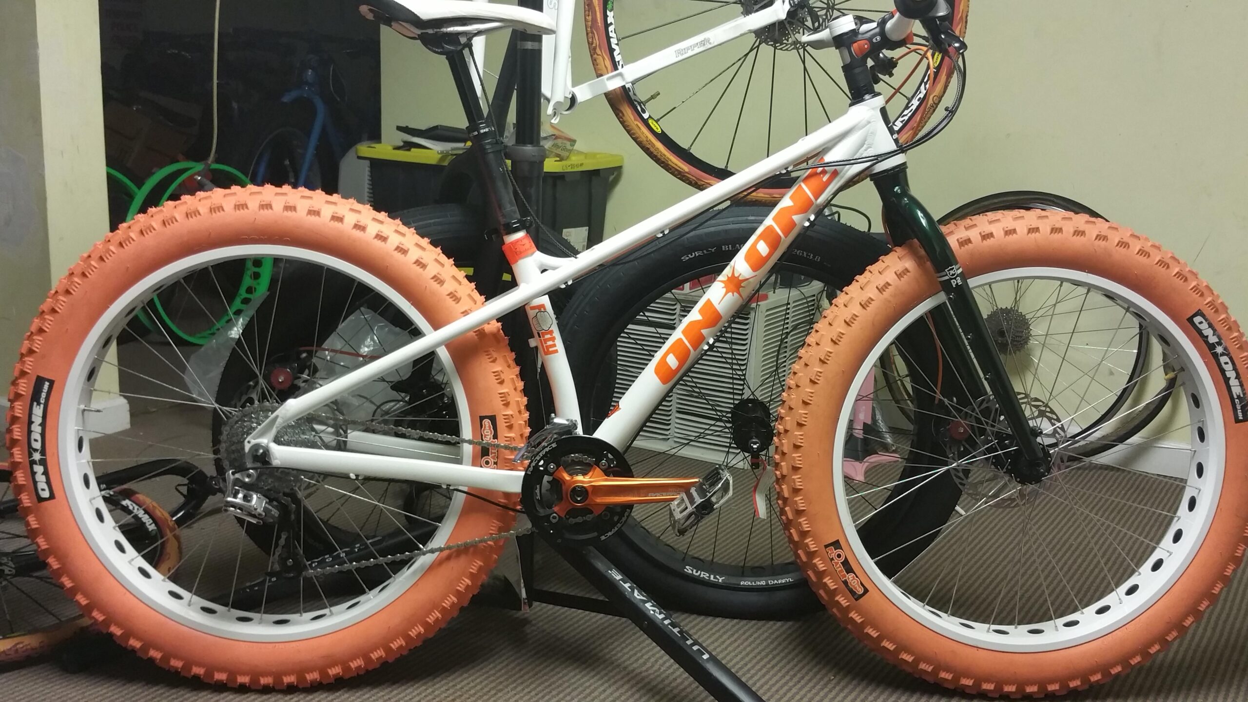 On-One Fatty: A fat tire mountain bike with distinctive orange tires and a white frame, positioned in a cluttered indoor space. The bike features prominent branding and a visible drivetrain. Other bicycles and items can be seen in the background.