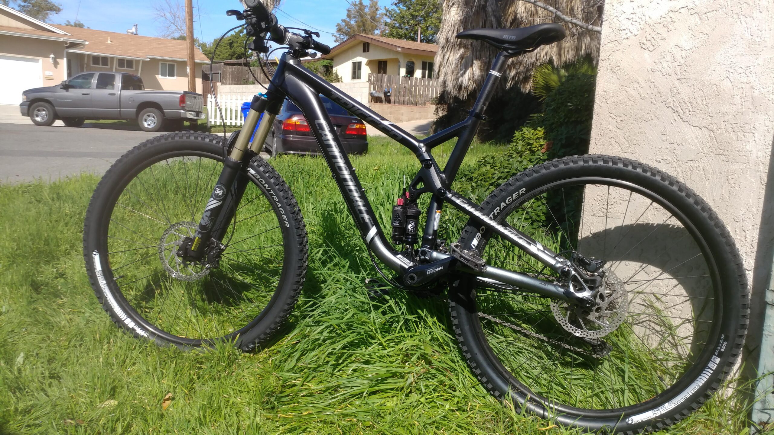 Cannondale Trigger: A black and silver mountain bike is resting on lush green grass. The bike features thick, knobby tires and a front suspension fork. In the background, a residential street can be seen with parked vehicles and houses. The scene is set on a sunny day.