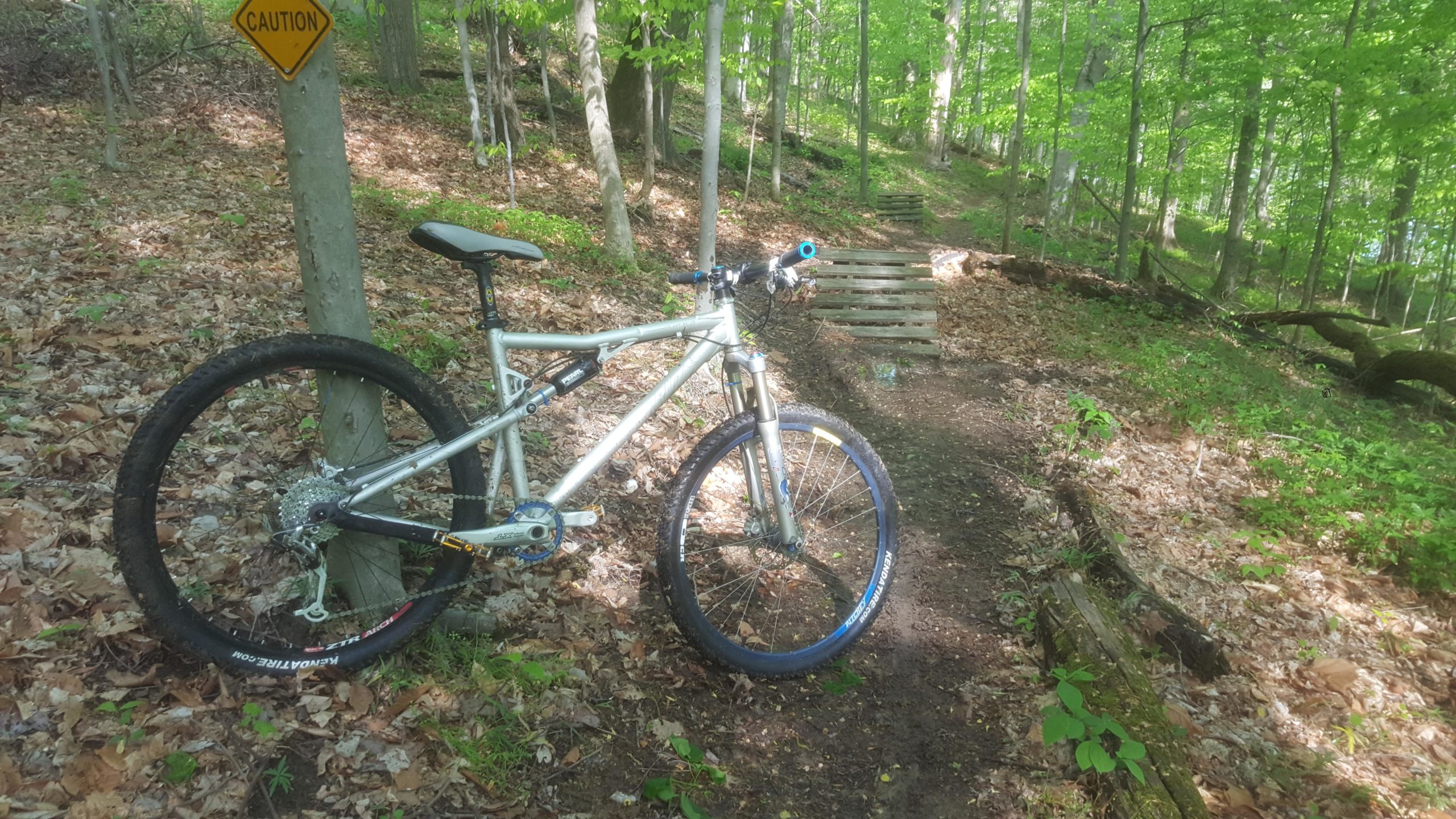 A mountain bike leaning against a tree on a dirt trail surrounded by green foliage. A caution sign is visible nearby, and wooden structures are seen in the distance along the path, indicating a natural, wooded area suitable for biking. Dillon State Park mountain bike trail.