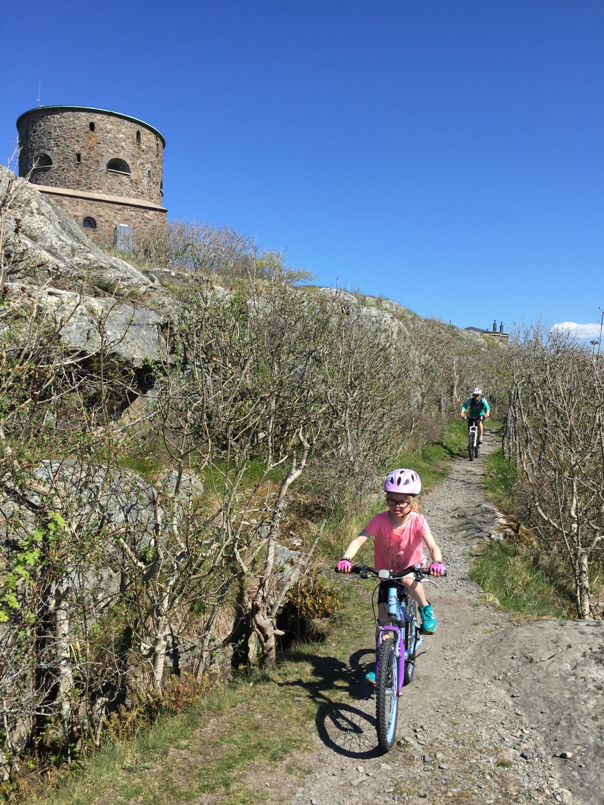 A young girl in a pink shirt and helmet rides a purple mountain bike on a trail surrounded by bushes, with a stone tower in the background against a clear blue sky. Another cyclist can be seen further along the path. Marstrand Island Trail System mountain bike trail.