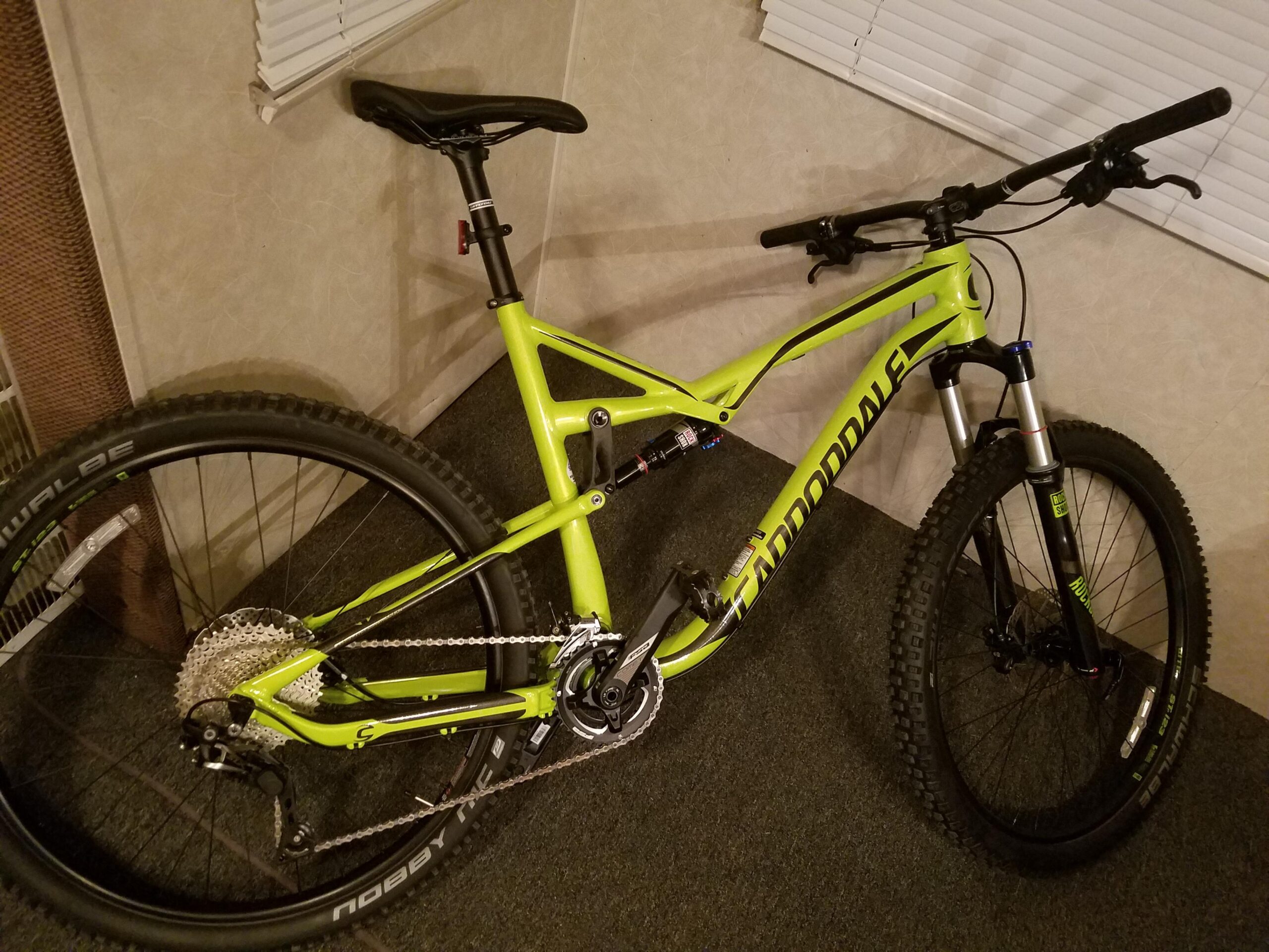 Cannondale Habit: A brightly colored green Cannondale mountain bike leaning against a wall with its wheels on a carpeted floor. The bike features wide tires, a visible gear system, and a front suspension fork, with handlebars positioned at a comfortable height for the rider. The background includes a window with blinds partially drawn.