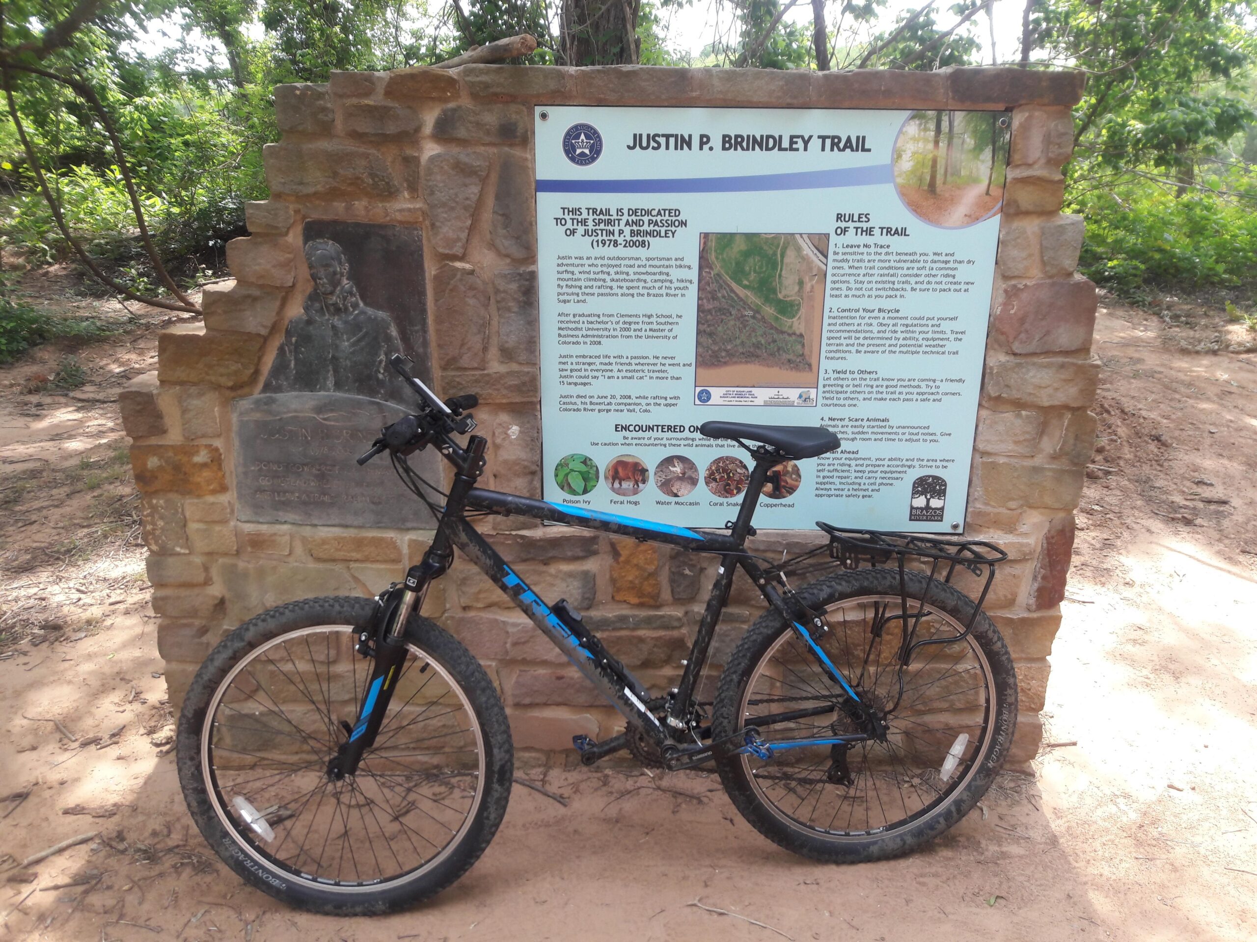 Trek 820: A mountain bike rests against a stone wall featuring a plaque and an informational sign about the Justin P. Brindley Trail. The sign includes rules for trail use and a dedication to Justin P. Brindley, with a description of the trail and its surroundings. Lush greenery is visible in the background, indicating a natural setting.