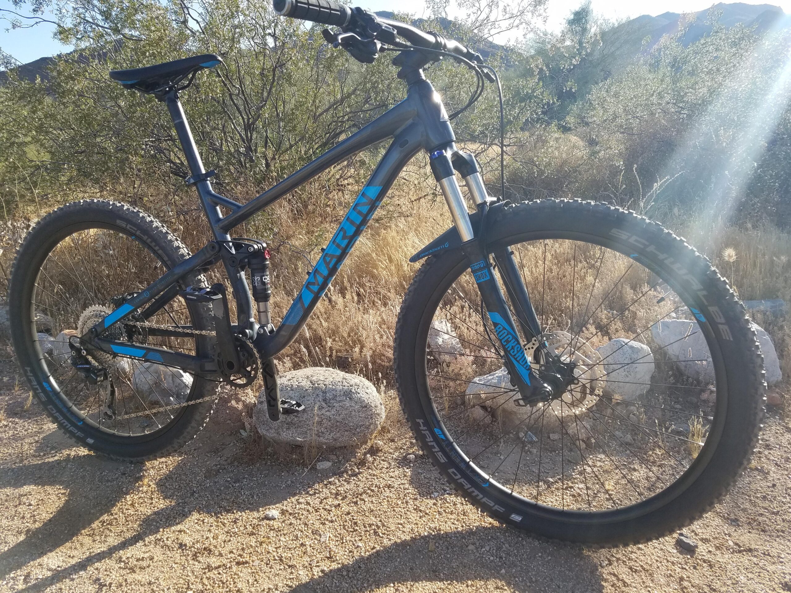 Marin Hawk Hill: A mountain bike with a black and blue frame is parked on a rocky surface surrounded by dry grass and sparse vegetation. Sunlight beams down, illuminating the bicycle, which features thick tires and a front suspension fork, indicating it's designed for trail riding. The background includes distant hills and a variety of plants.