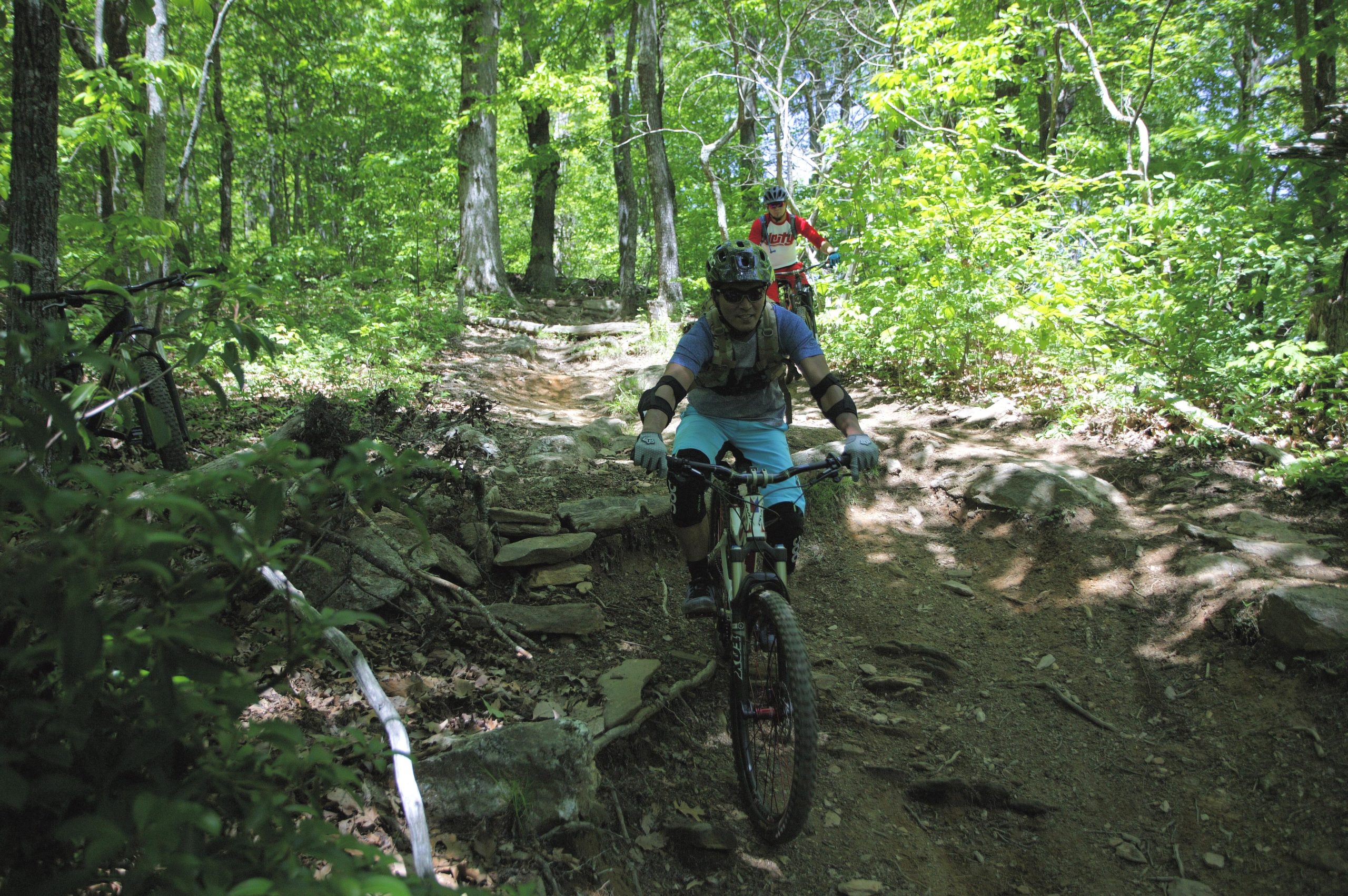 A person riding a mountain bike along a rocky trail in a lush green forest. The cyclist is wearing protective gear, including a helmet and gloves, while navigating through the terrain. In the background, another cyclist is visible, also riding on the trail. Sunlight filters through the trees, highlighting the vibrant foliage. Black Mountain mountain bike trail.