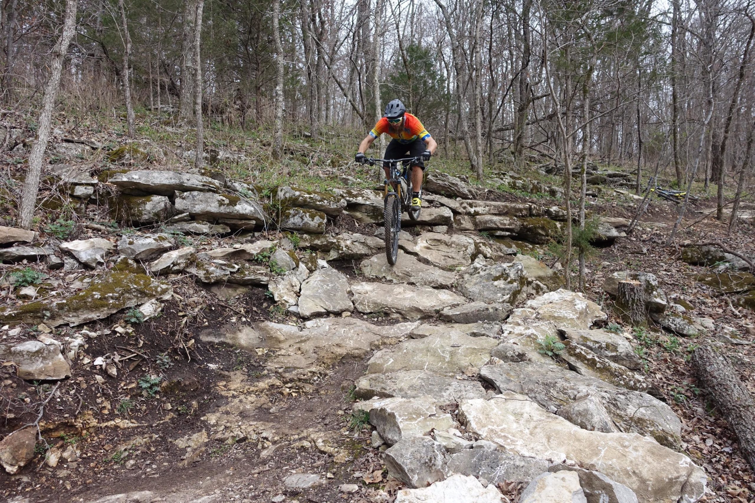 A mountain biker navigating a rocky trail in a wooded area, surrounded by trees and foliage. The rider is in an active position on the bike, maneuvering over large, uneven stone surfaces. Coler Mountain Bike Preserve mountain bike trail.
