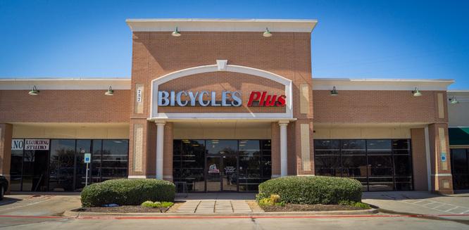 Front view of a brick storefront for "Bicycles Plus," featuring large signage in red and blue, with glass windows and green shrubbery lining the entrance. The sky is clear and blue, indicating a sunny day.