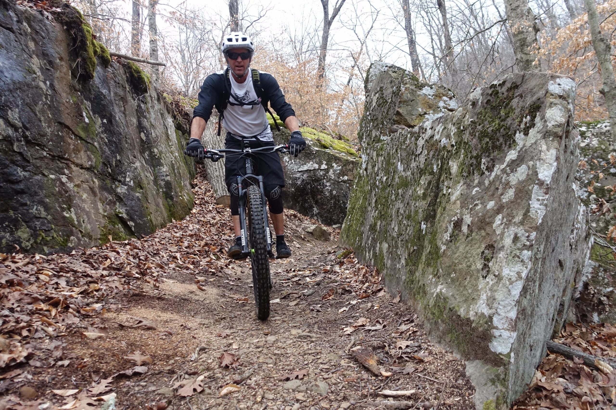 A mountain biker navigating a narrow trail surrounded by large rocks and autumn leaves, with trees in the background and a slightly overcast sky. The biker is wearing a helmet, sunglasses, and protective gear, and is focused on the path ahead. Upper Buffalo Headwaters Trail System mountain bike trail.