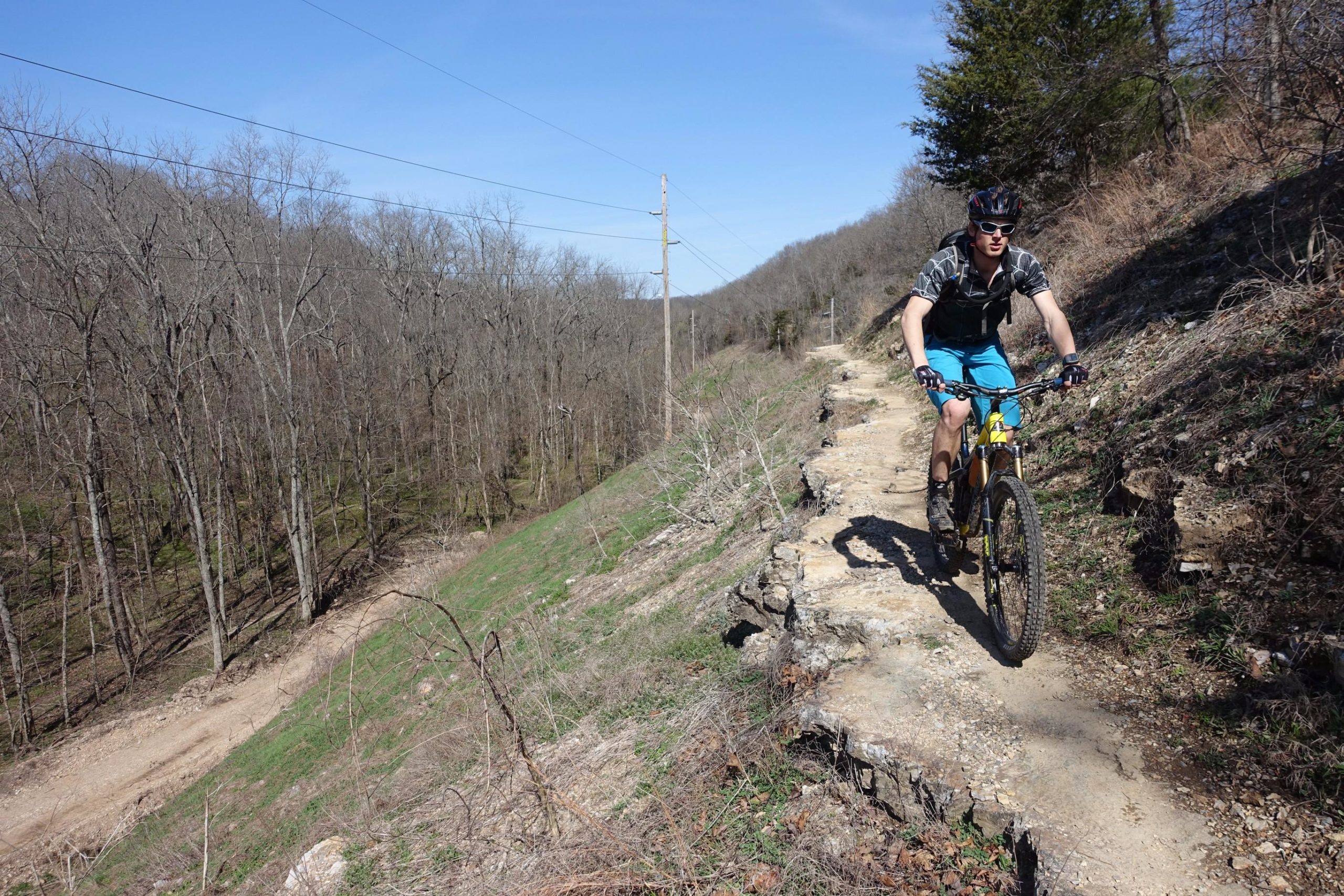 A person riding a mountain bike along a narrow dirt trail, surrounded by bare trees and green grass on a sunny day. Power lines stretch across the clear blue sky in the background. Back 40 mountain bike trail.