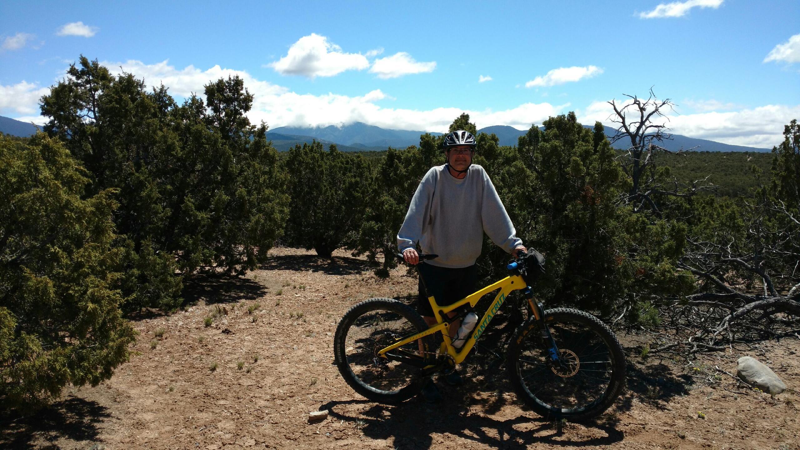 An individual wearing a helmet and a gray sweatshirt stands next to a yellow mountain bike on a dirt pathway surrounded by green shrubs and trees. In the background, there are mountains under a clear blue sky with a few clouds. La Tierra mountain bike trail.