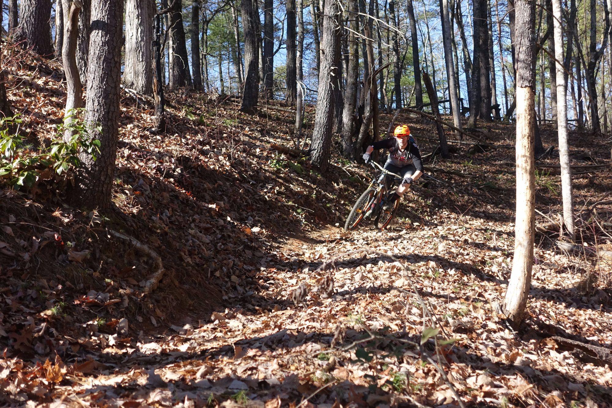 A mountain biker in a bright orange helmet is leaning into a turn on a leaf-covered trail in a forested area. Sunlight filters through the trees, casting shadows on the ground as the biker navigates a winding path. Bull / Jake Mountain mountain bike trail.