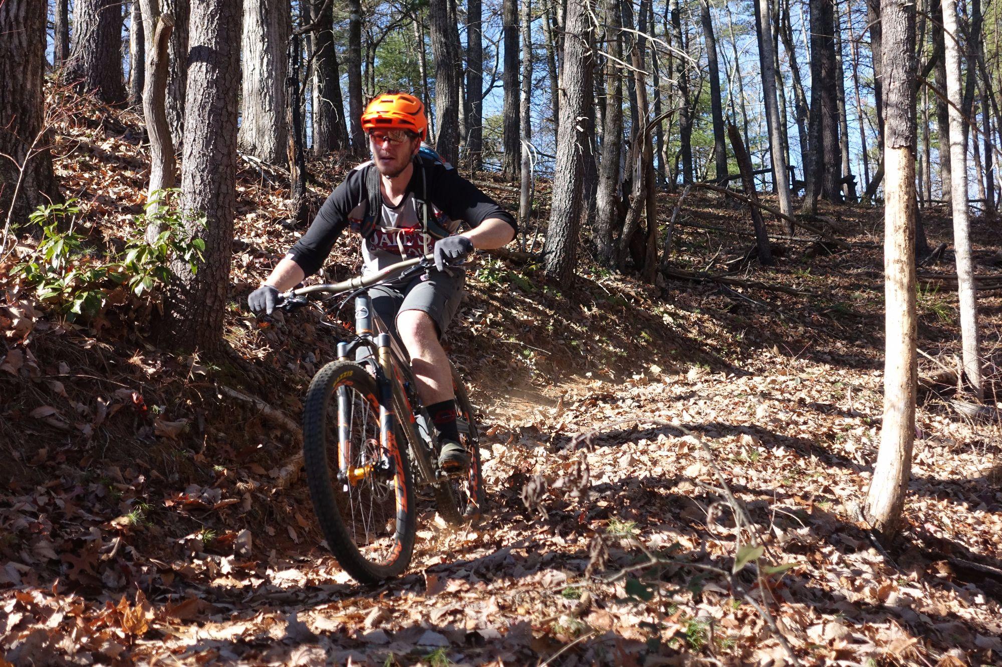A mountain biker riding through a forested trail covered in fallen leaves. The cyclist is wearing an orange helmet and a black and white shirt, focused on navigating the winding path surrounded by trees. Sunlight filters through the branches, creating a clear, bright day for biking. Bull / Jake Mountain mountain bike trail.