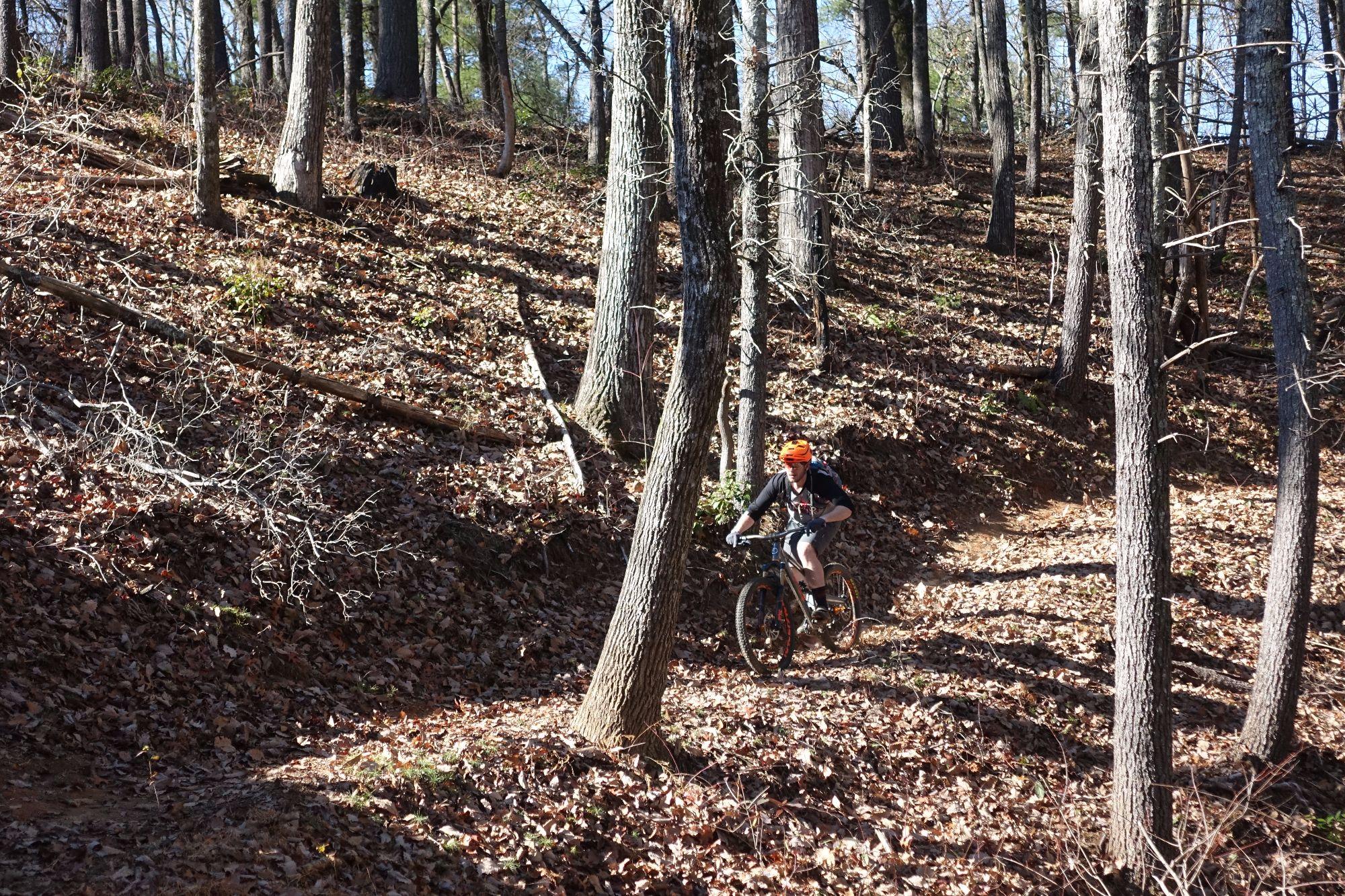 A person riding a mountain bike on a dirt trail through a wooded area during the fall, surrounded by trees and fallen leaves. The cyclist is wearing an orange helmet and casual attire. Bull / Jake Mountain mountain bike trail.