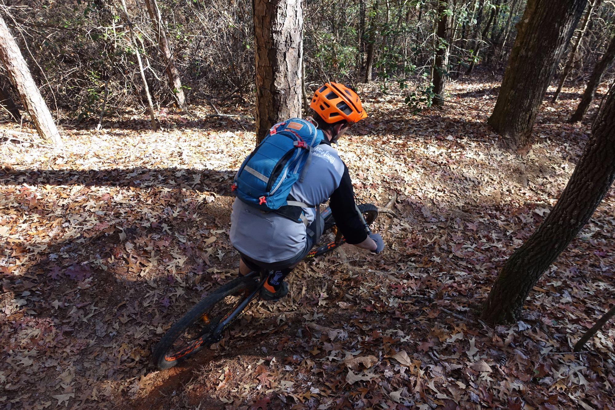 A mountain biker navigates a wooded trail covered with fallen leaves. The rider, wearing an orange helmet and a gray cycling outfit, leans into the turn while descending the path. A blue backpack is secured to their back, and tall trees and underbrush frame the scene, creating a natural outdoor setting. Bull / Jake Mountain mountain bike trail.