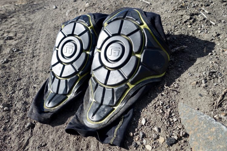 A pair of black knee pads with textured protective shells resting on a rocky, dirt surface. The pads feature yellow accents and are designed for impact protection in sports or outdoor activities.