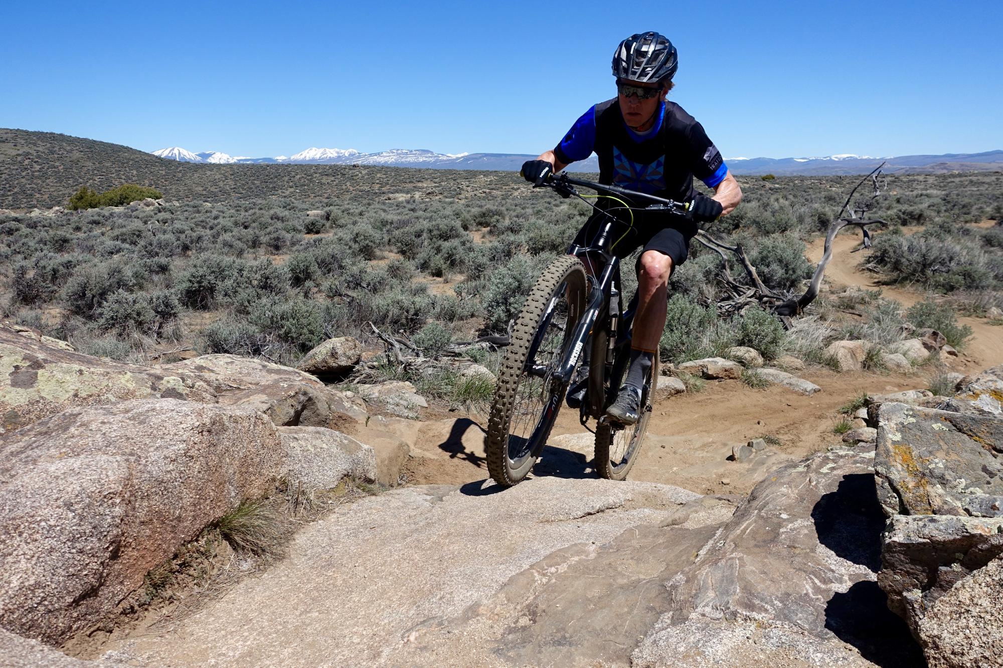 A person riding a mountain bike on a rocky trail in a vast outdoor landscape. The scenery features shrubs and distant mountains under a clear blue sky. The cyclist is focused, navigating over a rocky surface with one wheel lifted off the ground. Hartman Rocks mountain bike trail.