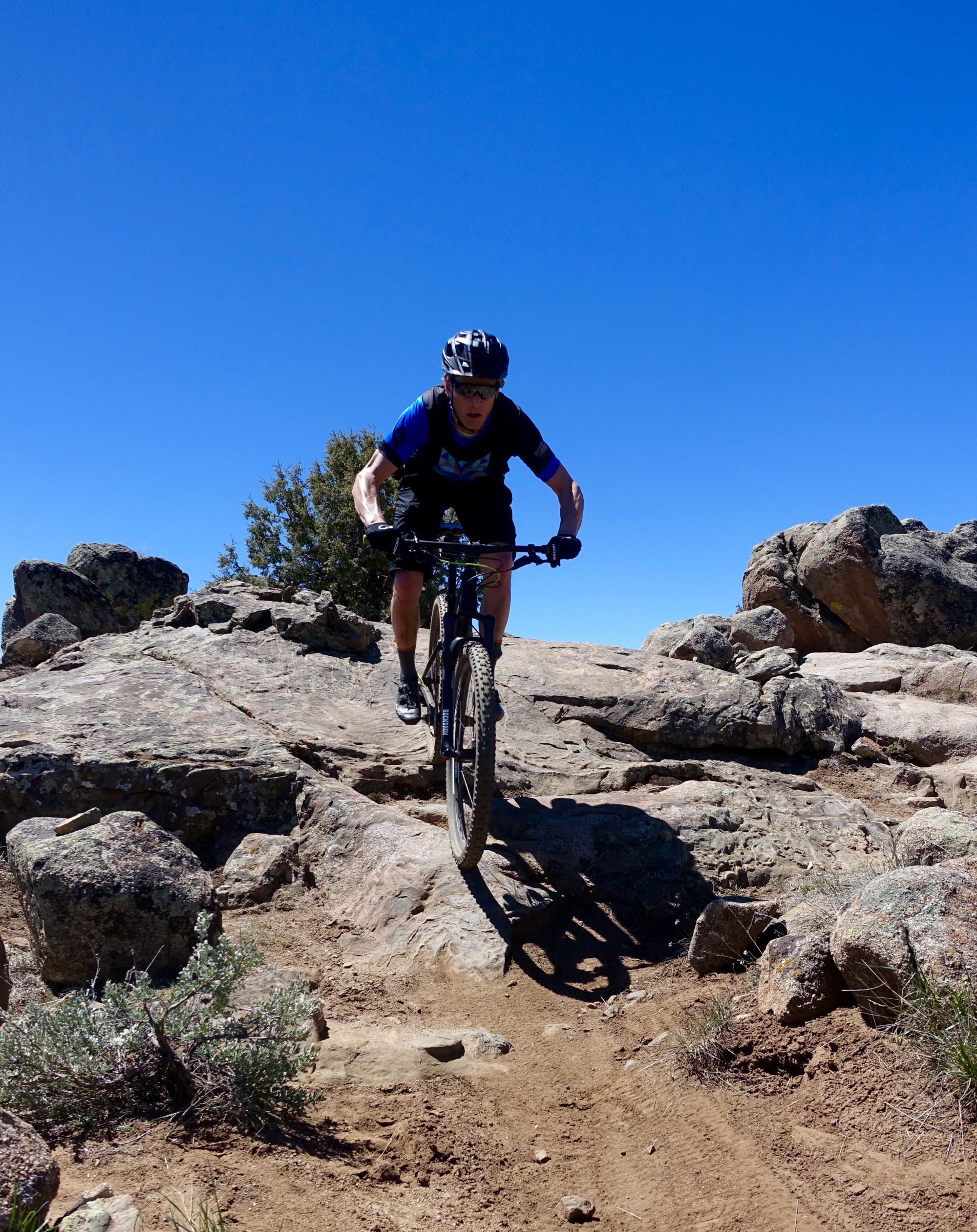 A mountain biker navigating rocky terrain under a clear blue sky, demonstrating an action shot as they ride over uneven rocks. The surrounding landscape features boulders and sparse vegetation. Hartman Rocks mountain bike trail.