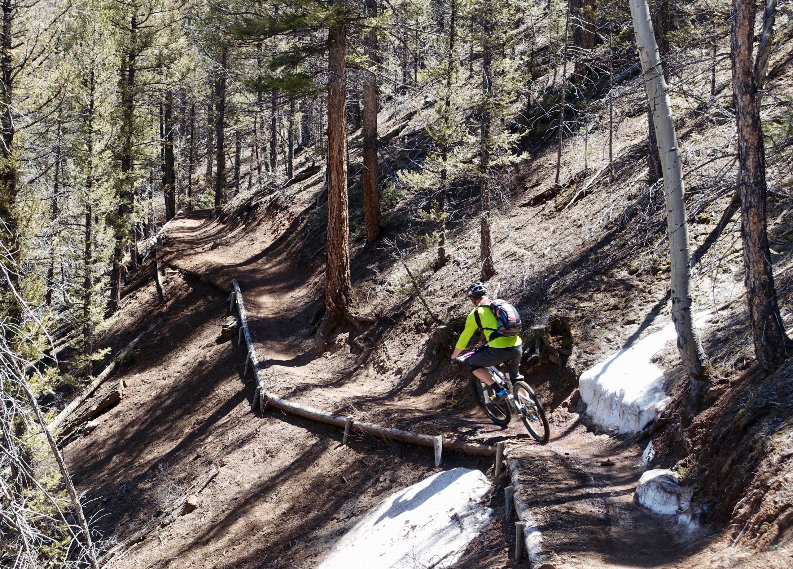 A mountain biker riding on a dirt trail through a pine forest, with sunlight filtering through the trees. The path winds alongside some patches of snow, and wooden barriers are visible along the trail. Rainbow Trail: Silver Creek to Hwy 285 mountain bike trail.