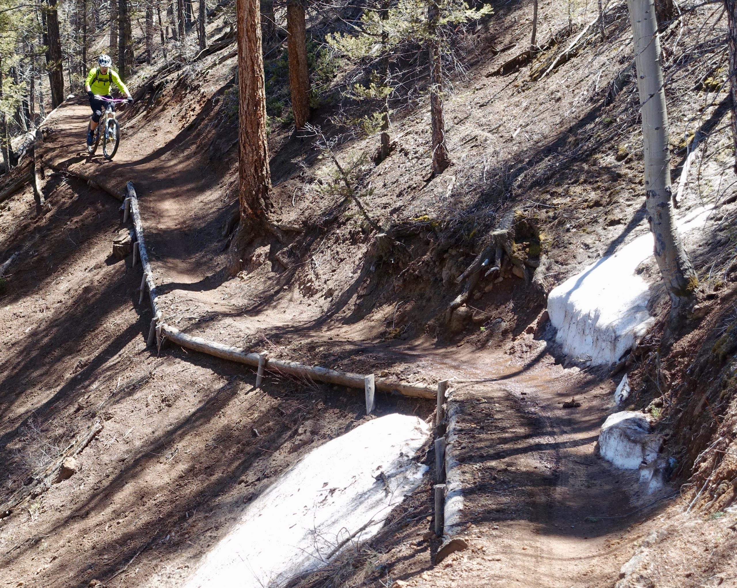 A mountain biker wearing a bright yellow shirt rides along a dirt trail winding through a forest. The path is bordered by wooden logs and features areas of remaining snow. Sunlight filters through the trees, creating dappled shadows on the ground. Rainbow Trail: Silver Creek to Hwy 285 mountain bike trail.