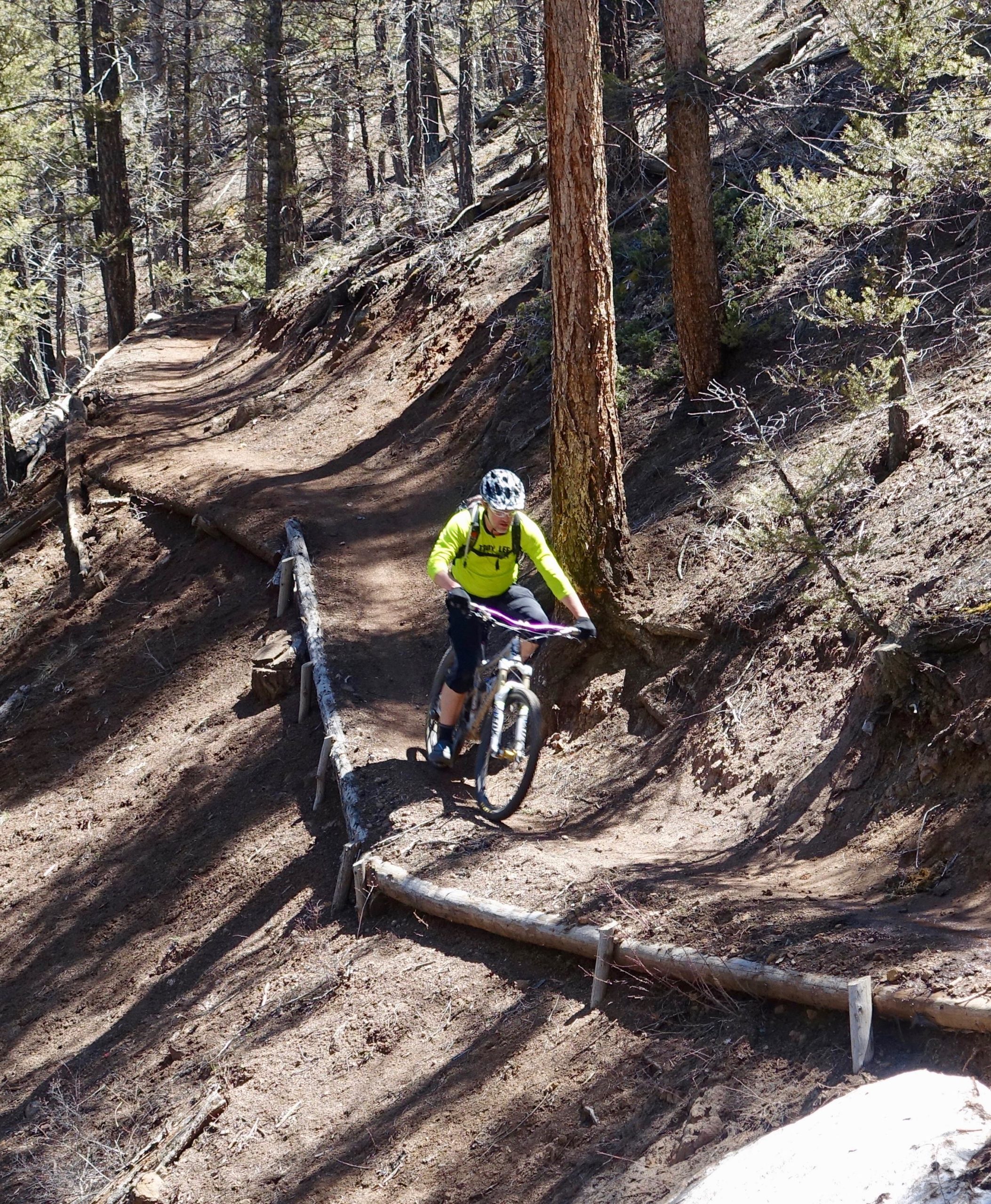 A mountain biker wearing a bright green long-sleeve shirt and helmet navigates a winding dirt trail in a wooded area, surrounded by tall trees and natural terrain. Rainbow Trail: Silver Creek to Hwy 285 mountain bike trail.