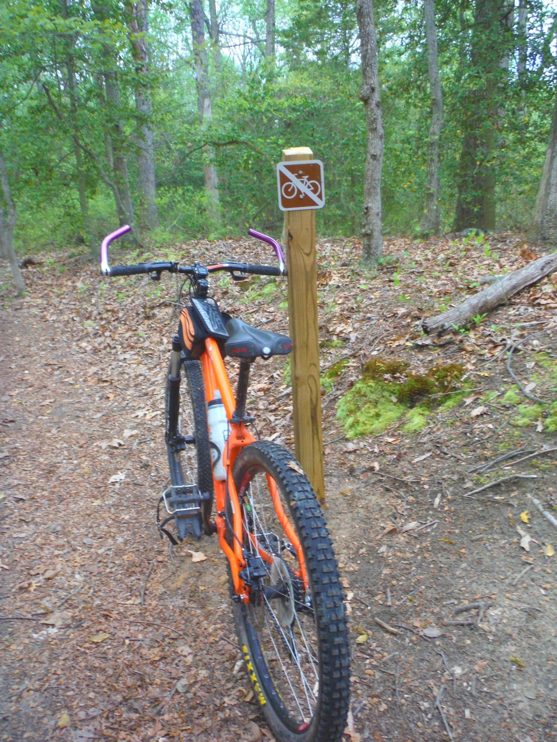 An orange mountain bike leaning against a wooden post with a "no biking" sign, surrounded by forested terrain covered in leaves and moss. Rancocas State Park - Westampton mountain bike trail.